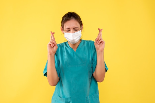 Вид спереди женщина-врач в медицинской рубашке и маске со скрещенными пальцами на желтом столе пандемическая больница цвет covid - вирус здоровья медик