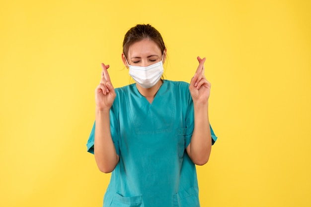 Front view female doctor in medical shirt and mask with crossed fingers on yellow desk pandemic hospital color covid- health virus medic