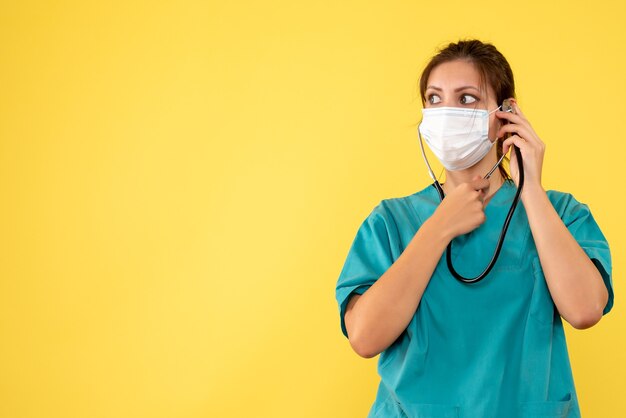 Front view female doctor in medical shirt and mask with stethoscope on yellow background