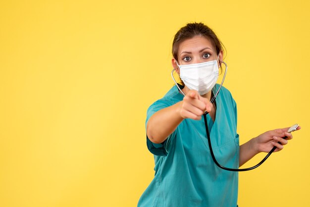 Front view female doctor in medical shirt and mask with stethoscope on yellow background