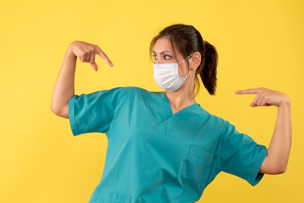 Front view female doctor in medical shirt with sterile mask pointing at herself on yellow background