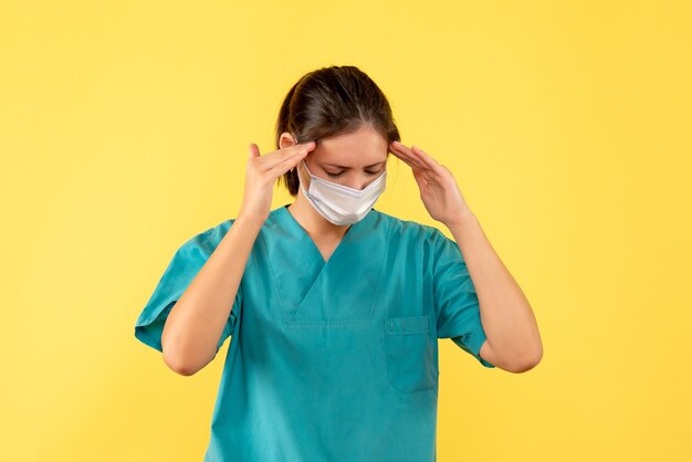 黄色の背景に頭痛に苦しんでいる滅菌マスクと医療シャツの正面図の女性医師