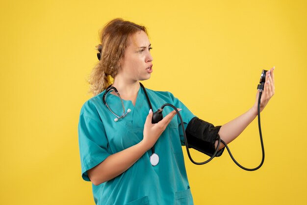 Front view of female doctor in medical suit checking her pressure on yellow wall