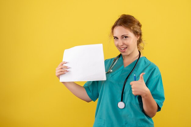 Front view of female doctor in medical suit holding analysis on a yellow wall
