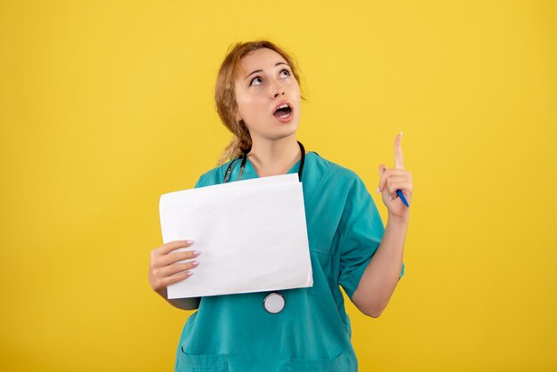 Front view of female doctor in medical suit holding analysis on yellow wall