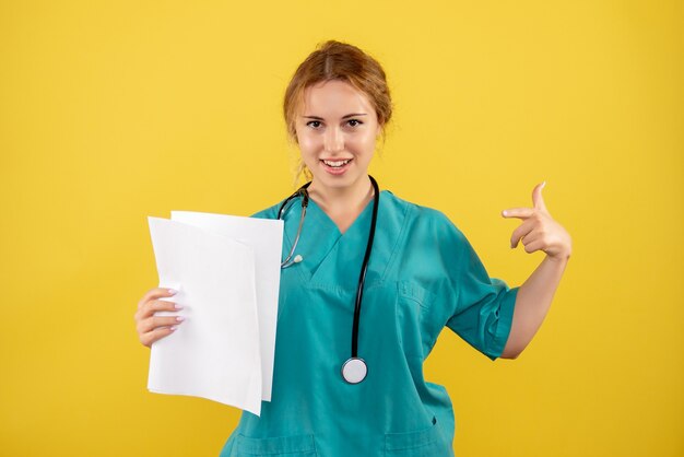 Вид спереди женщины-врача в медицинском костюме, держащей анализ на желтой стене