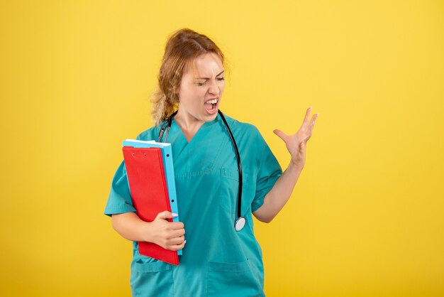 Вид спереди женщины-врача в медицинском костюме, держащей различные анализы на желтой стене