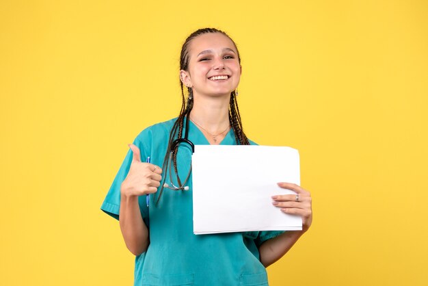 Front view of female doctor in medical suit holding papers on a yellow wall