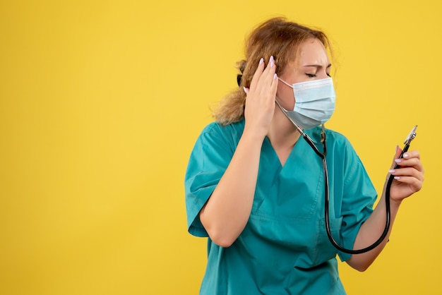 Front view of female doctor in medical suit and mask with stethoscope on a yellow wall