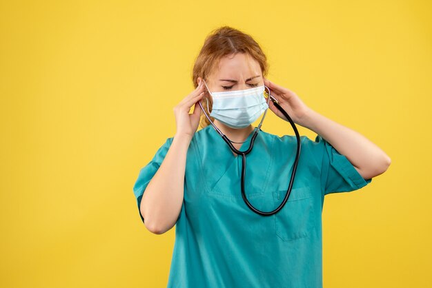 Front view of female doctor in medical suit and mask with stethoscope on yellow wall