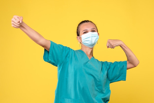 Вид спереди женщины-врача в медицинском костюме и стерильной маске на желтой стене