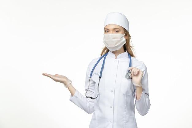 Front view female doctor in medical suit wearing mask due to coronavirus holding a stethoscope on light-white wall virus pandemic covid- illness