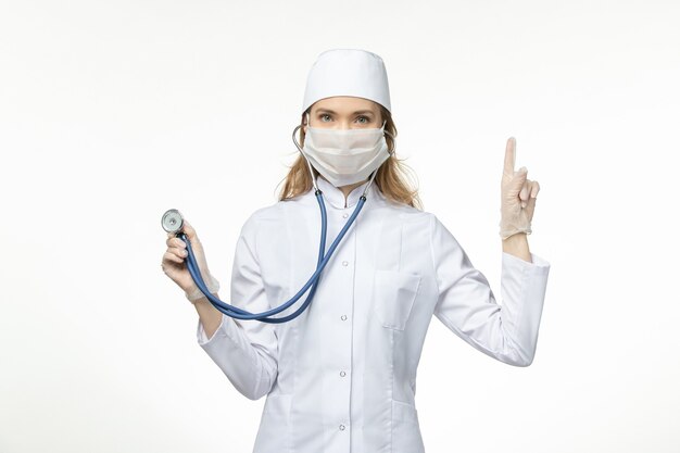 Front view female doctor in medical suit wearing mask due to coronavirus holding stethoscope on light white wall virus pandemic covid- illness