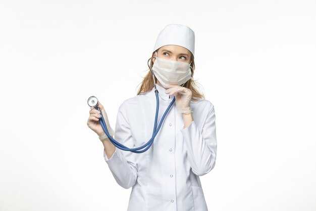 Front view female doctor in medical suit wearing mask due to coronavirus holding stethoscope thinking on the white wall virus pandemic covid- illness