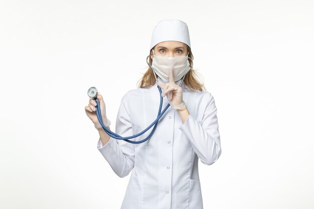 Front view female doctor in medical suit wearing mask due to coronavirus holding stethoscope on the white wall pandemic covid- illness