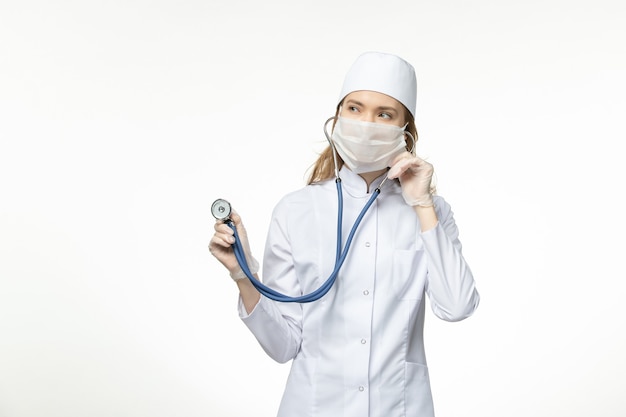 Вид спереди женщина-врач в медицинском костюме в маске из-за коронавируса, держащая стетоскоп на белой стене, пандемическое заболевание ковидом