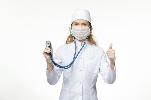 Front view female doctor in medical suit wearing mask due to coronavirus holding stethoscope on white wall virus pandemic covid- illness