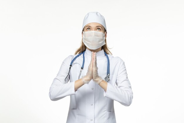 Вид спереди женщина-врач в медицинском костюме в маске из-за коронавируса молится о болезни светло-белого стола, заболевании, вызванном пандемическим вирусом