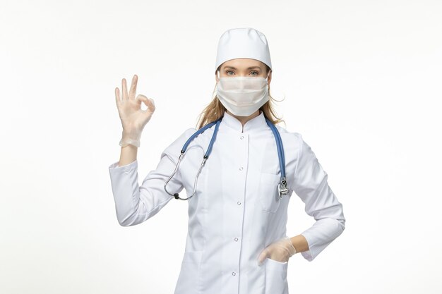 Front view female doctor in medical suit wearing mask due to coronavirus on the white wall disease covid- pandemic disease illness