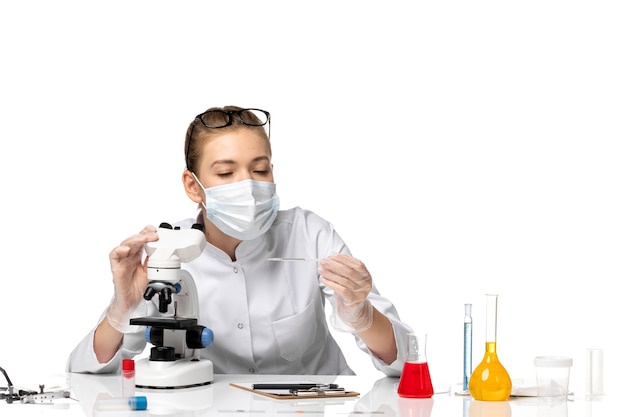 Front view female doctor in medical suit wearing mask due to covid using microscope on the white space