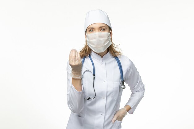 Front view female doctor in medical suit wearing mask and gloves due to coronavirus on light white wall pandemic covid- disease virus illness