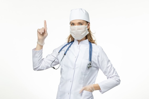 Front view female doctor in medical suit wearing mask and gloves due to coronavirus on white wall covid- pandemic disease virus illness