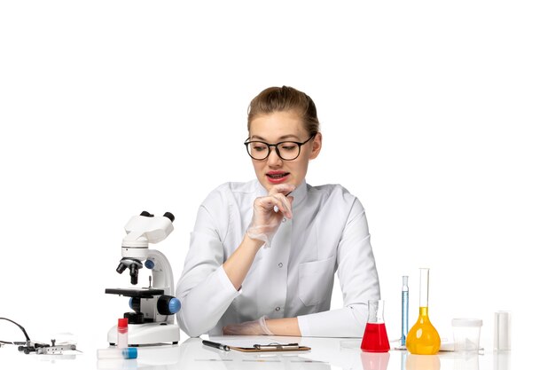 Front view female doctor in medical suit with gloves sitting with solutions on white desk