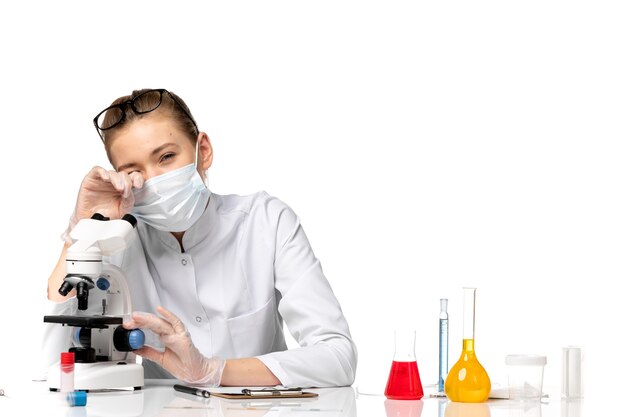 Front view female doctor in medical suit with mask due to covid using microscope on light white space