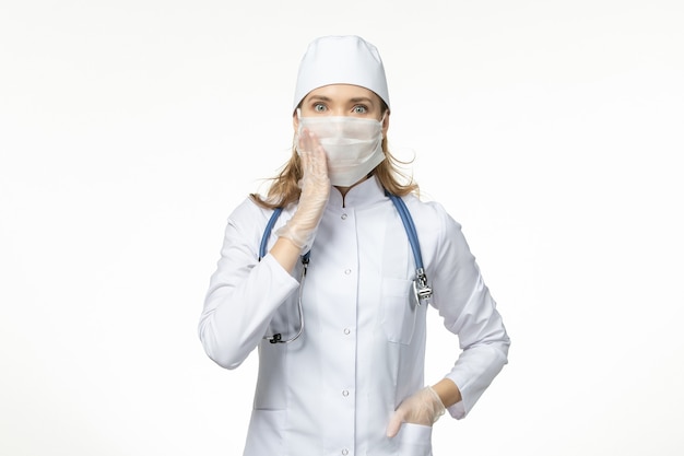 Medico femminile di vista frontale in vestito medico con maschera e guanti a causa del coronavirus sul virus covid pandemico della malattia del muro bianco chiaro