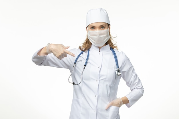 Front view female doctor in medical suit with mask and gloves due to coronavirus on the light-white wall disease pandemic covid- virus