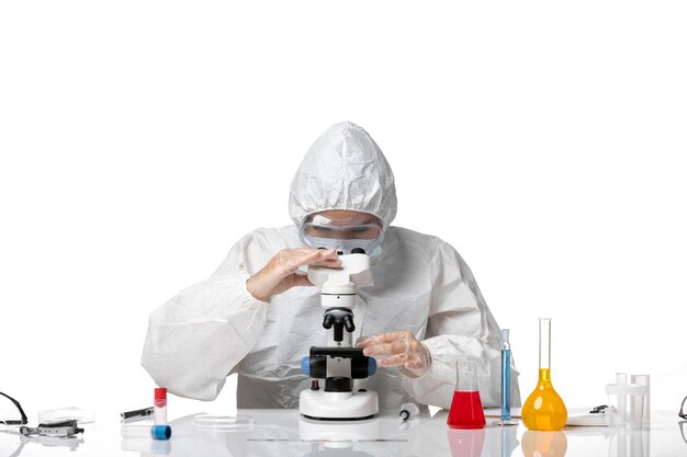 Front view female doctor in protective suit with mask due to covid using microscope on light white background virus pandemic health covid