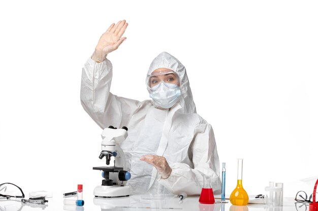 白い背景のウイルスパンデミックスプラッシュcovidに手を振っている顕微鏡を使用してcovidによるマスク付きの防護服を着た正面図の女性医師-