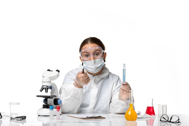 Вид спереди женщина-врач в защитном костюме и с маской, держащая синий раствор и пишущая заметки на белом фоне, пандемический вирус covid, коронавирус