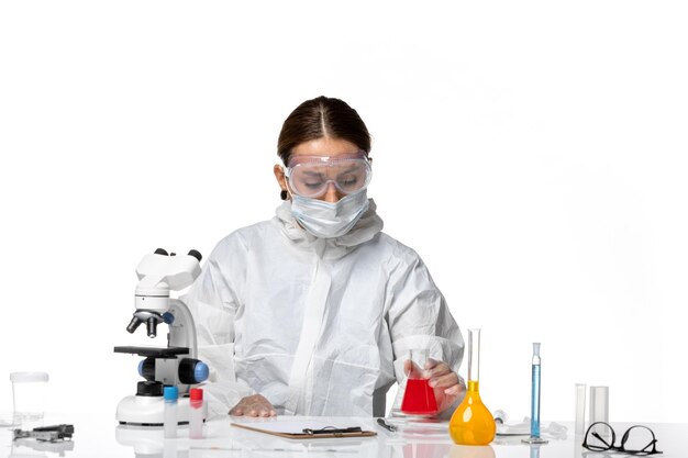 Front view female doctor in protective suit and with mask holding flask with red solution on light white background covid virus pandemic coronavirus