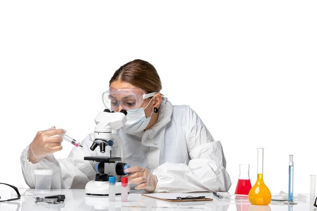 Front view female doctor in protective suit and with mask using microscope on light white background health covid virus pandemic coronavirus