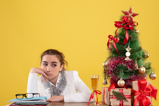 Вид спереди женщина-врач сидит вокруг рождественских подарков и скучает елке на желтом фоне