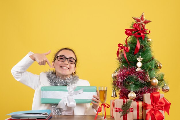 Вид спереди женщина-врач сидит вокруг рождественских подарков и елки с подарком на желтом фоне