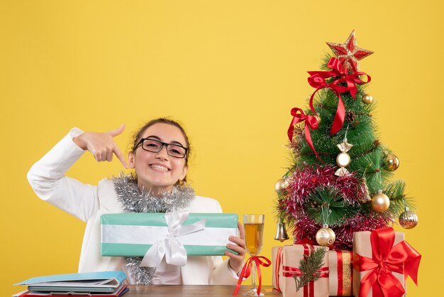 Front view female doctor sitting around christmas presents and tree holding gift on yellow background