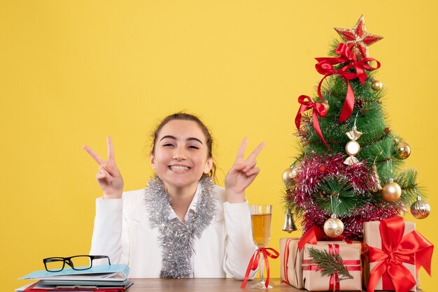 Front view female doctor sitting around christmas presents and tree posing with smile on yellow background