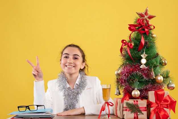 Front view female doctor sitting around christmas presents and tree smiling on yellow background