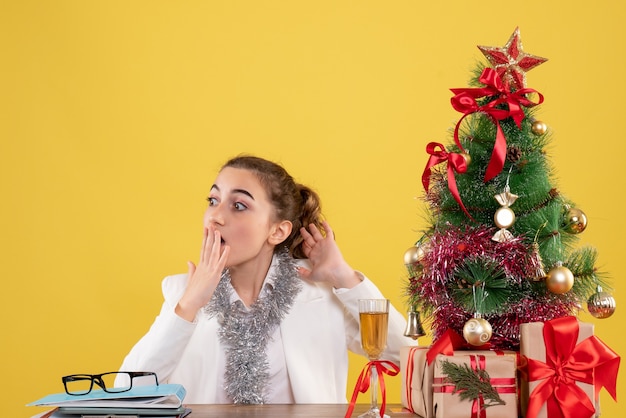 Medico femminile di vista frontale che si siede intorno ai regali di natale e all'albero su fondo giallo