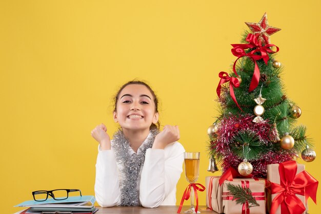 Вид спереди женщина-врач сидит вокруг рождественских подарков и елки на желтом фоне