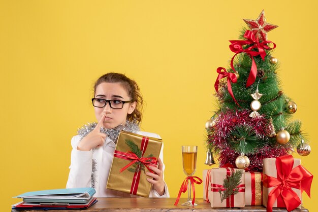クリスマスプレゼントと黄色の背景の木の周りに座っている正面図の女性医師