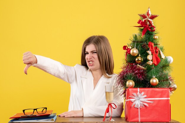 彼女のテーブルの前に座っている正面図の女性医師は、クリスマスツリーとギフトボックスと黄色の背景に不満を持っています