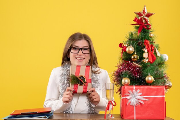 Front view female doctor sitting in front of her table holding present on yellow background with christmas tree and gift boxes