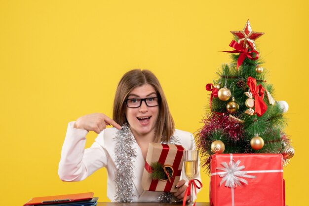クリスマスツリーとギフトボックスと黄色の背景にプレゼントを保持している彼女のテーブルの前に座っている正面図の女性医師
