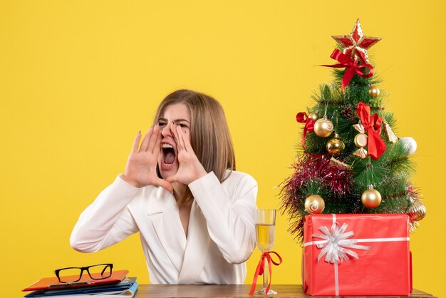 Front view female doctor sitting in front of her table loudly calling on yellow background with christmas tree and gift boxes