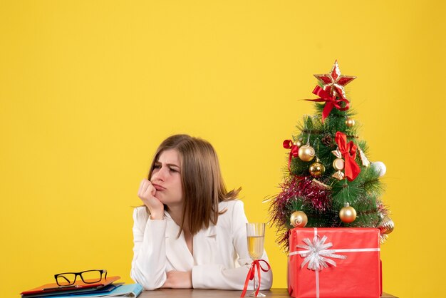 彼女のテーブルの前に座っている正面図の女性医師は、クリスマスツリーとギフトボックスで黄色の背景を強調