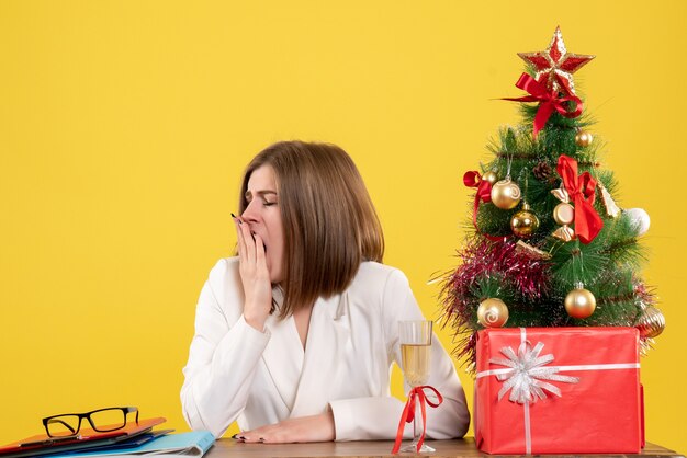 Вид спереди женщина-врач сидит перед своим столом, зевая на желтом фоне с рождественской елкой и подарочными коробками