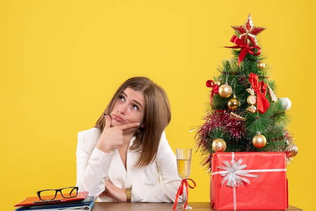 Front view female doctor sitting in front of her table on yellow background with christmas tree and gift boxes